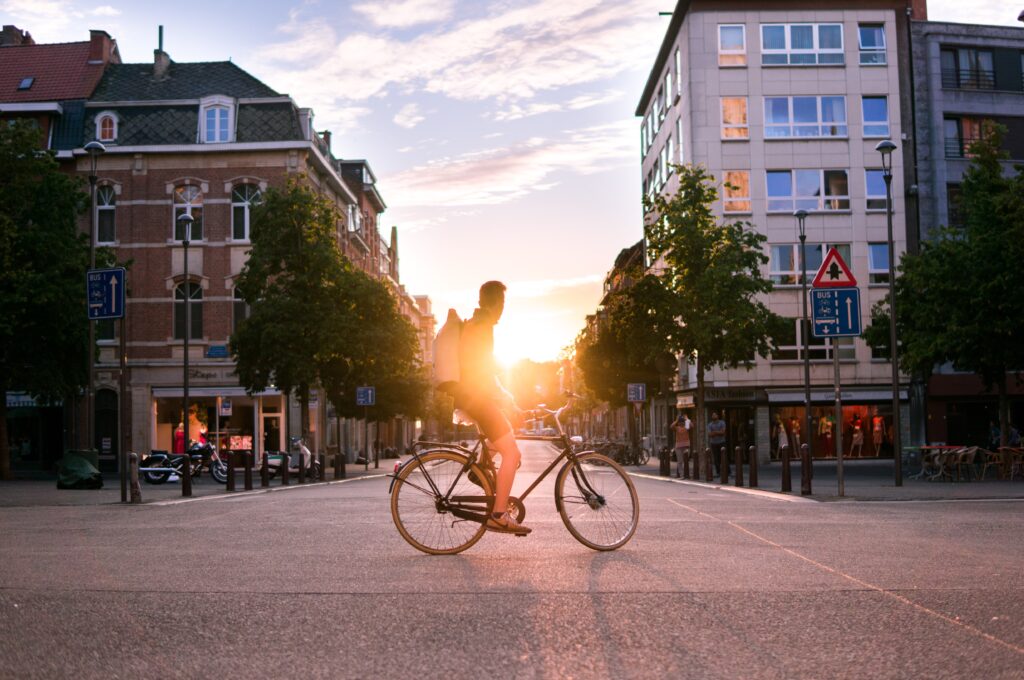 Eine Person steht mit einem Fahrrad in einer Fußgängerzone. IM Hintergrund geht die Sonne unter.