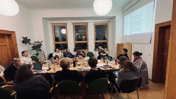 Viele Menschen sitzen um einen Tisch herum und diskutieren. Auf die Wand ist ein Text projiziert.